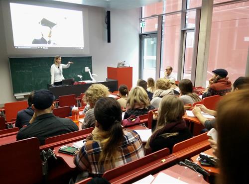 A Group of students is sitting in a lecture hall with red chairs. In the front a Young woman is giving a presentation.